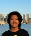 A young tan asian man with shoulder-length dark brown hair wearing a black t-shirt smiles in front of a body of water with a city in the background.